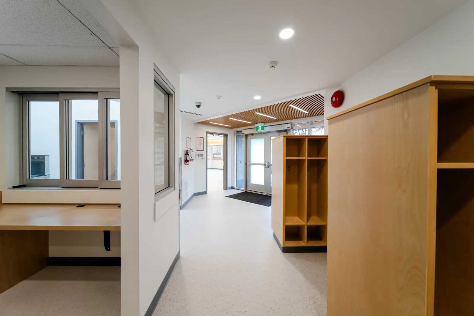 Childcare entrance area with wood cubbies, a staff office window and automatic entry doors under a slatted wood ceiling