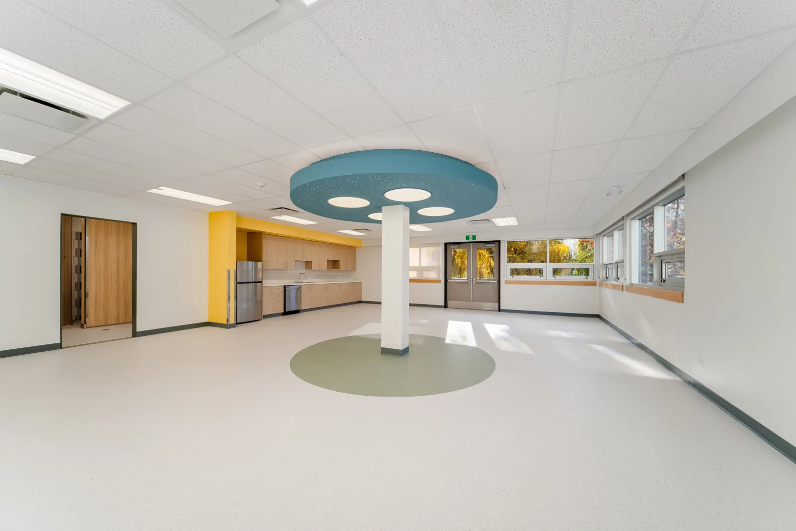 Open childcare classroom with a round blue acoustic ceiling feature, a green floor inset, a yellow-accented kitchenette and large windows