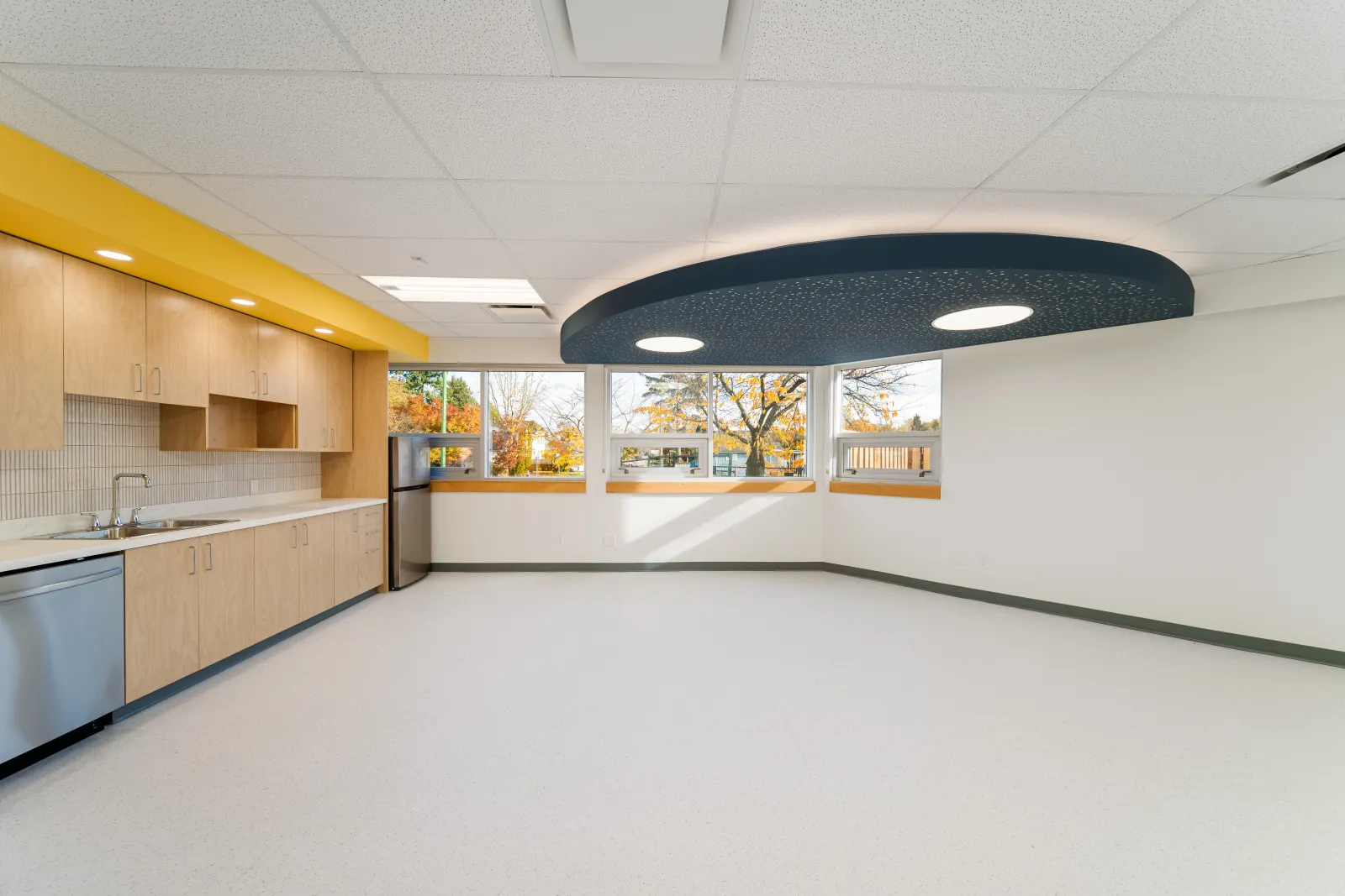 Bright classroom with wood cabinetry, a yellow bulkhead, a dark blue acoustic ceiling cloud and a row of windows