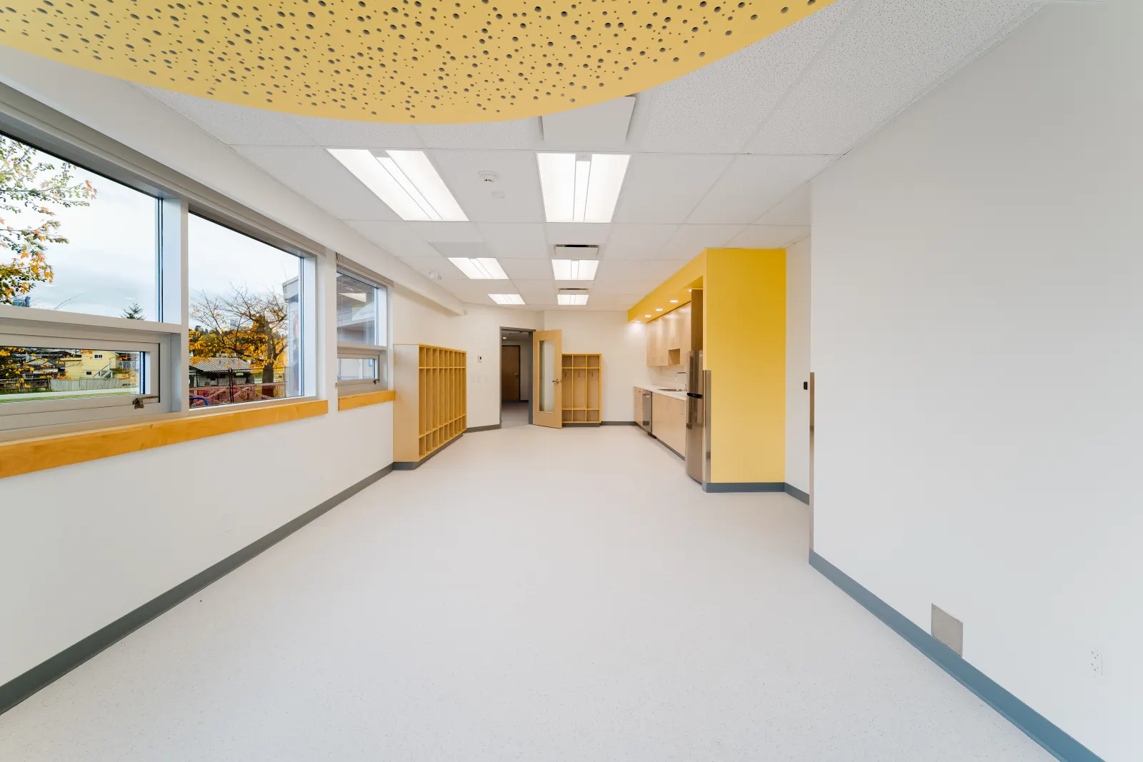 Long classroom with windows along one side, a perforated yellow ceiling feature, wood cubbies and a kitchenette at the far end
