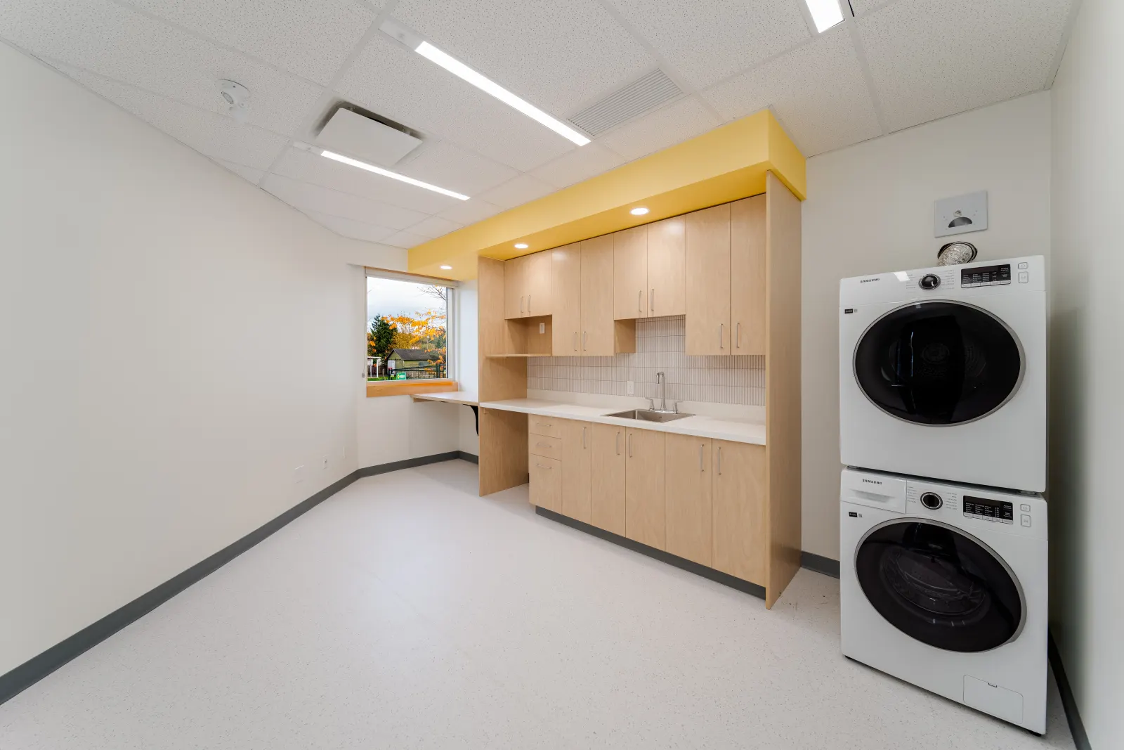 Room with a wood cabinet kitchenette under a yellow bulkhead, a window and a stacked washer and dryer