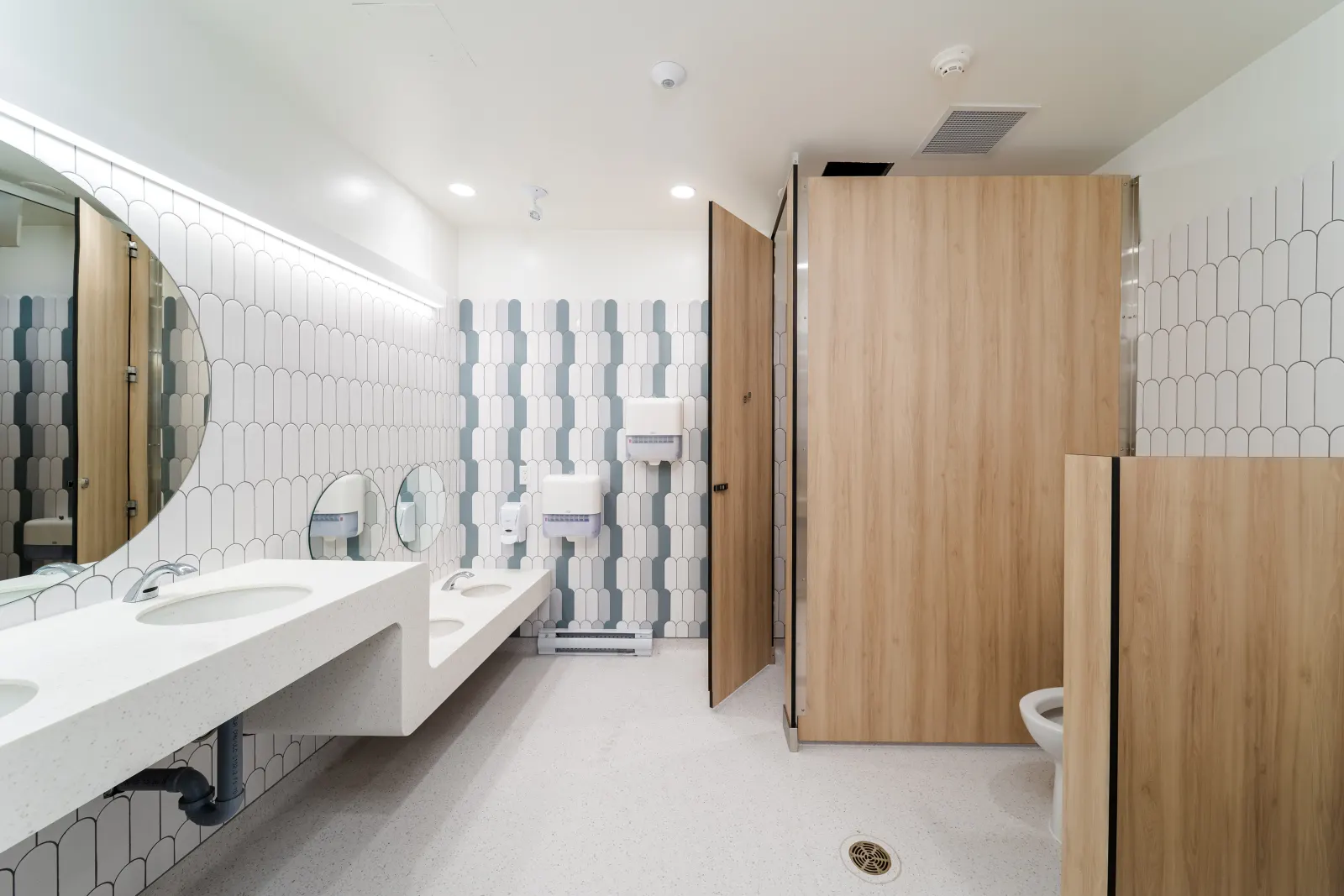 Washroom with white and green scalloped tile, a solid-surface counter with sinks at two heights, round mirrors and wood-grain stall partitions