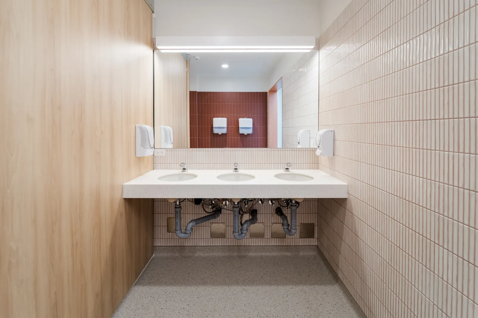Washroom vanity with three sinks on a solid-surface counter, pink vertical tile and a wood-grain wall