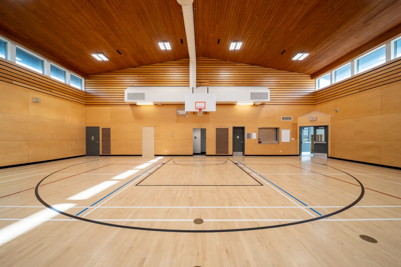 Community centre gym with maple sport flooring, a basketball hoop, wood-panelled walls and a wood ceiling